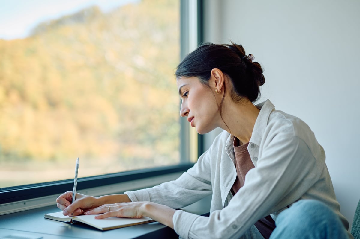 Young woman journaling thoughts in notebook while traveling by train, looking out window at scenery