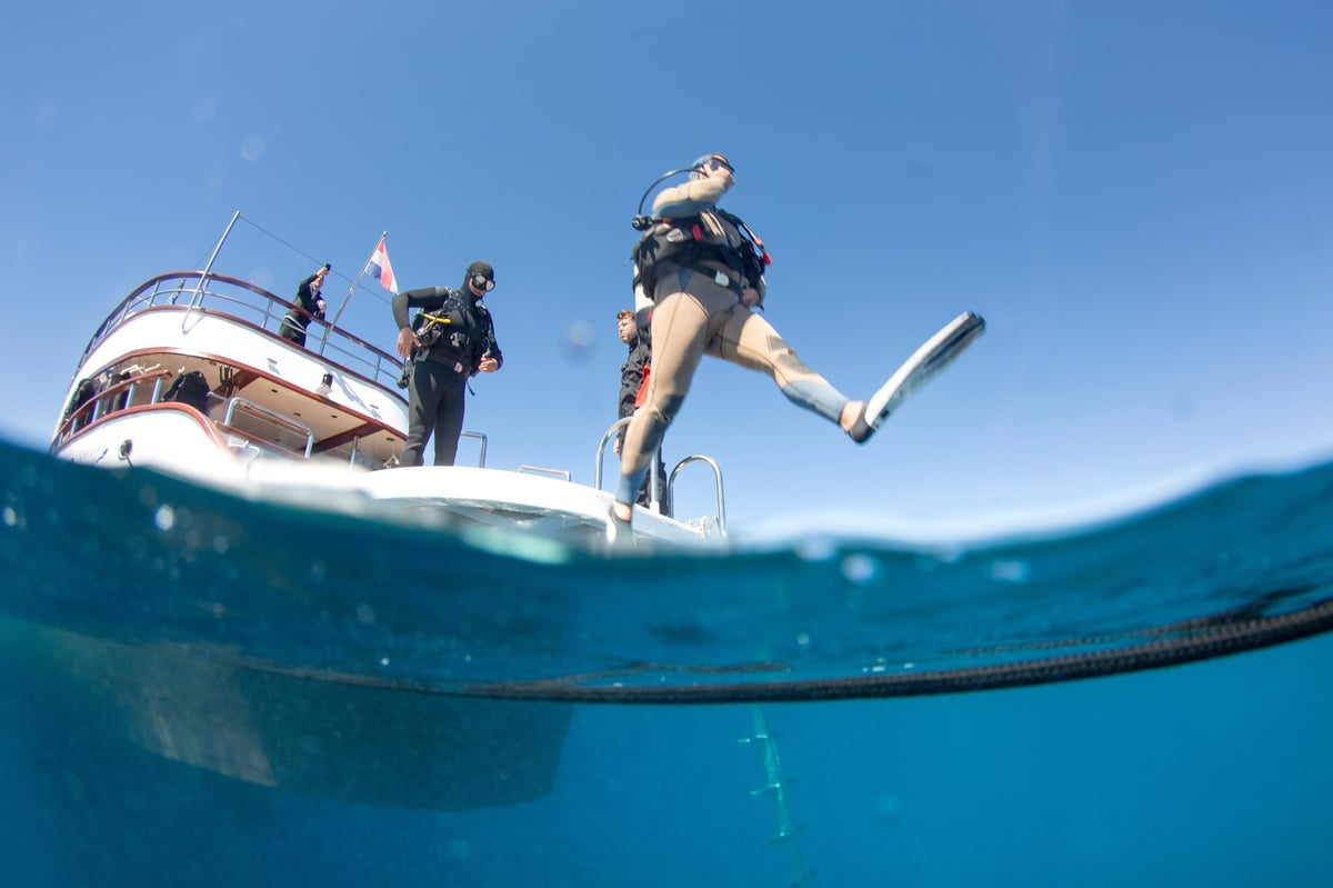 Divers are getting ready to explore underwater. One diver is jumping into the water while others prepare on the boat. Clear blue sea surrounds the scene under bright sunlight.