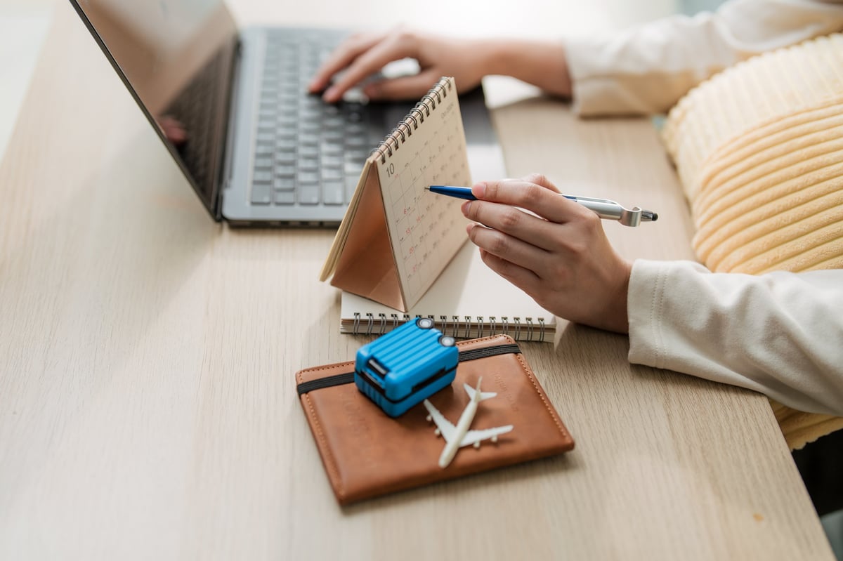 Conceptual image of a woman planning a business trip or summer vacation, booking flight tickets online with a laptop, passport, calendar, and miniature airplane on a wooden desk.